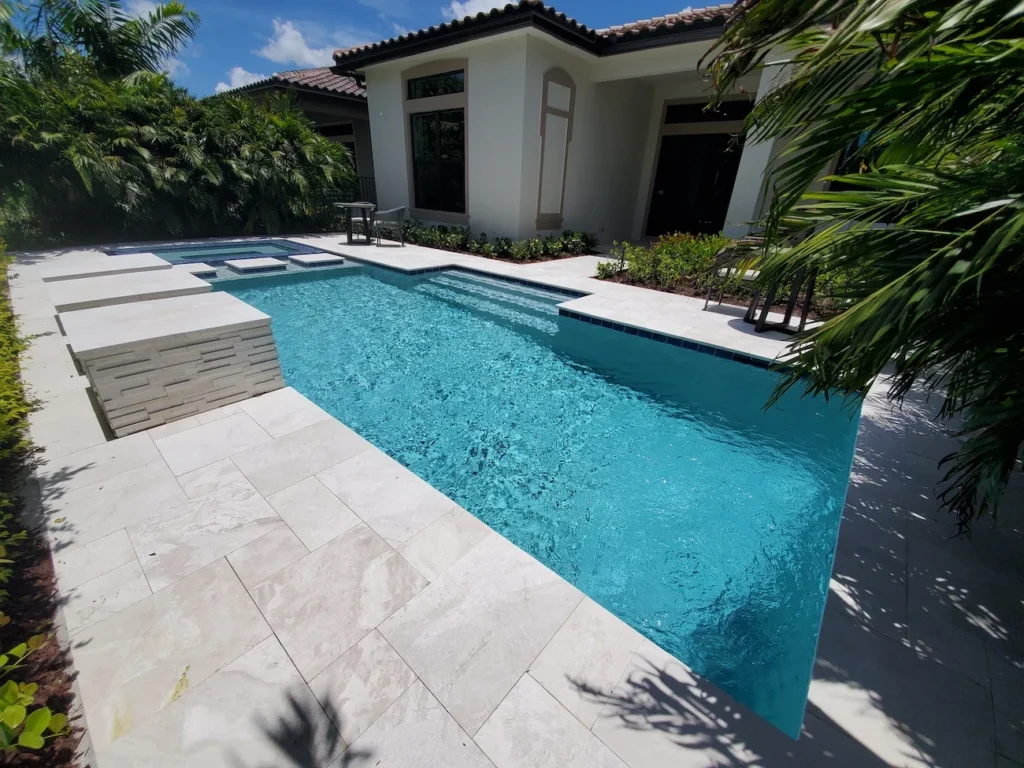 A rectangular backyard pool with a raised spa and stone accent wall beside a travertine paver deck.