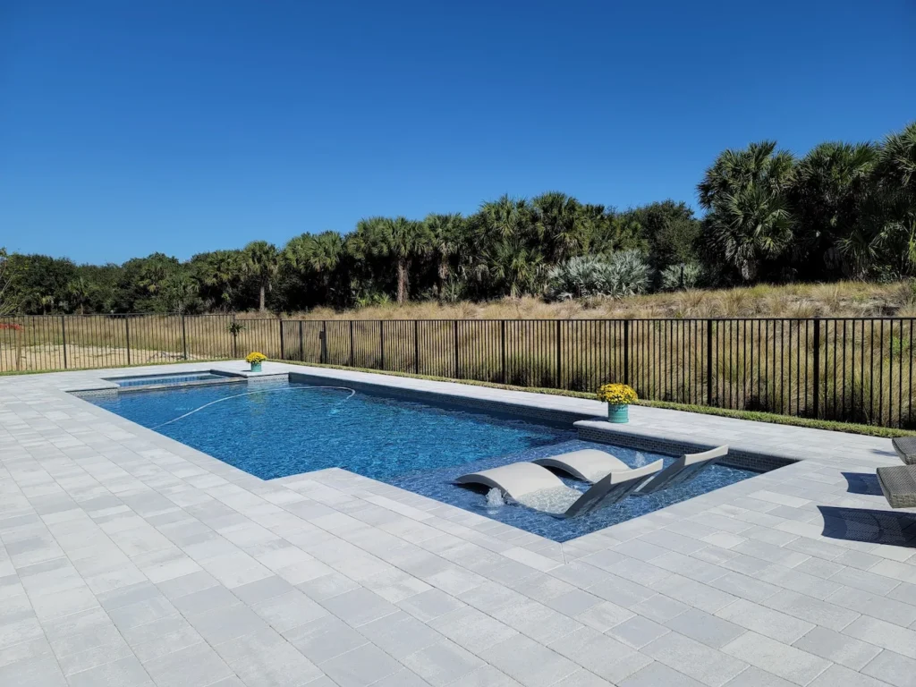 A rectangular backyard pool with a raised spa and two in-pool loungers with bubbling jets, surrounded by a paver deck and a rural tree line.