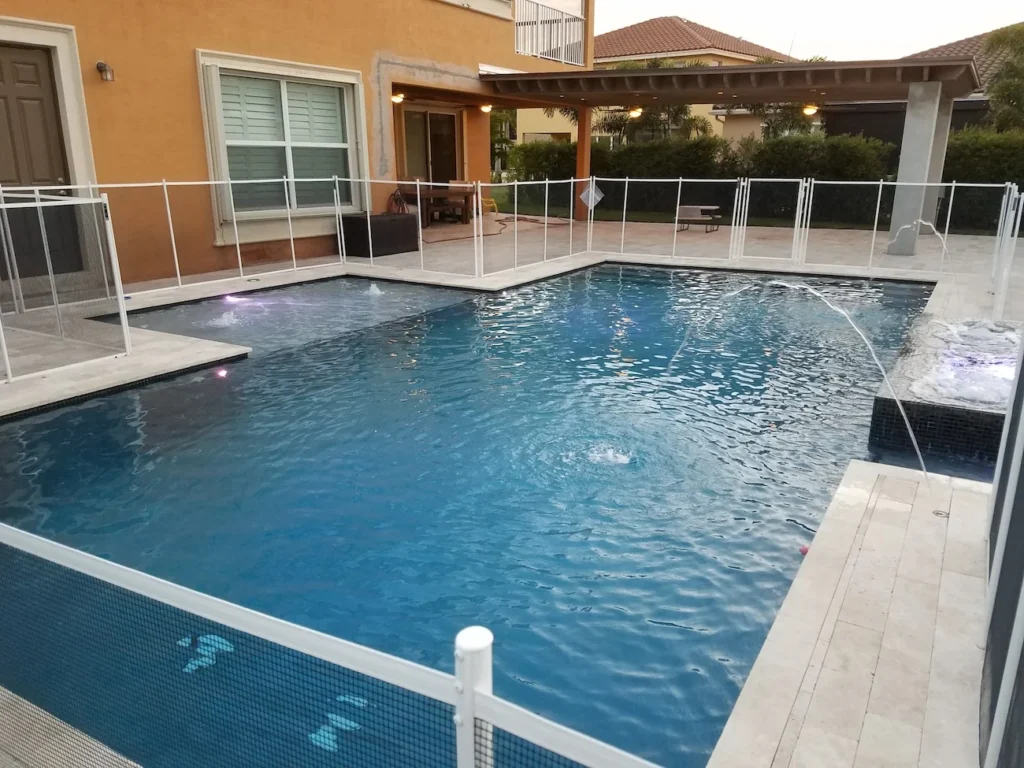 An L-shaped backyard pool with deck jets and a child safety fence at dusk.