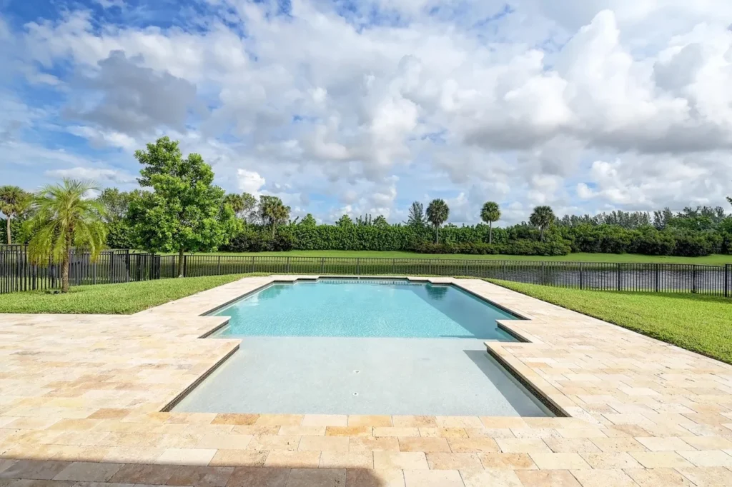 A rectangular pool with a shallow sun shelf, travertine deck, and open lawn behind a black metal fence.