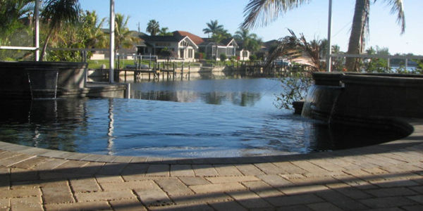 An infinity-edge pool with paver decking overlooking a residential canal with docked homes beyond.