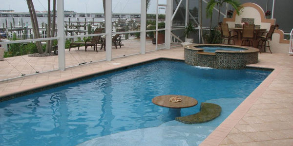 An L-shaped pool with a raised spillover spa, in-pool table, and paver deck inside a screened enclosure overlooking a marina.