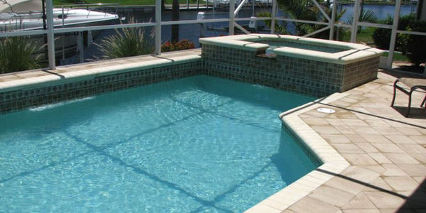 A rectangular pool with a raised tiled spa in a screened enclosure overlooking a canal with docked boats.