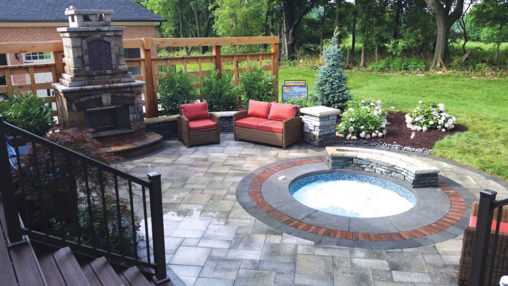 A round raised spa with blue tile and brick trim set into a paver patio with an outdoor fireplace and seating nearby.