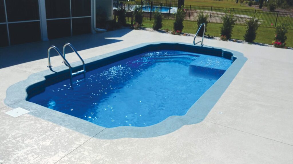 A small rectangular fiberglass pool with a raised sun shelf and steel handrails on a stamped concrete patio.