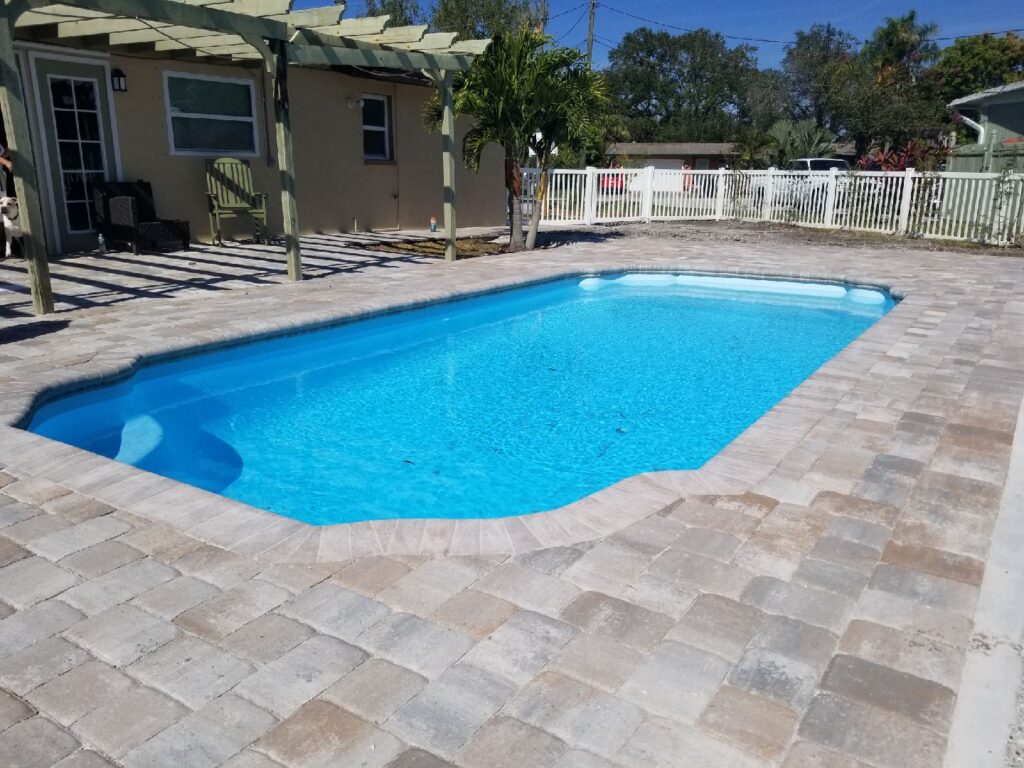 A rectangular fiberglass pool with a paver patio and pergola-covered lanai in a suburban backyard.