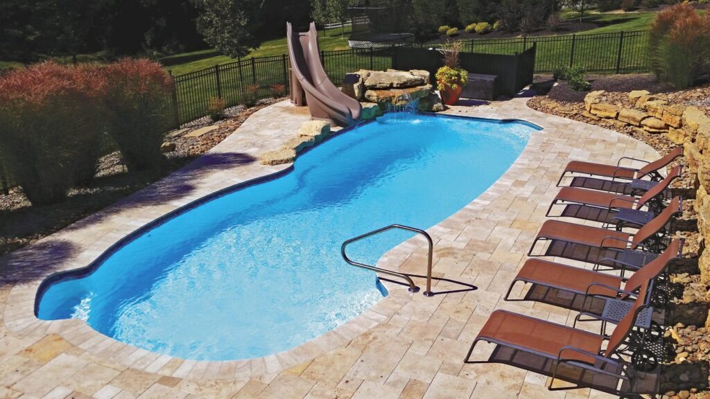 Freeform fiberglass pool with a waterslide, rock waterfall feature, and travertine paver deck lined with lounge chairs.