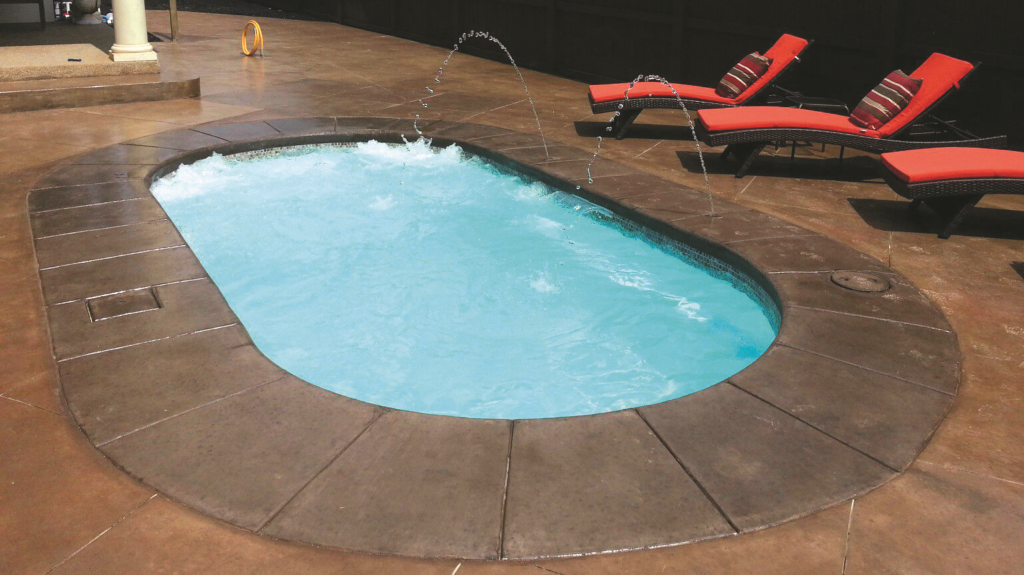A small oval fiberglass plunge pool with bubbling jets, surrounded by a stamped concrete patio and red lounge chairs, photographed at night.