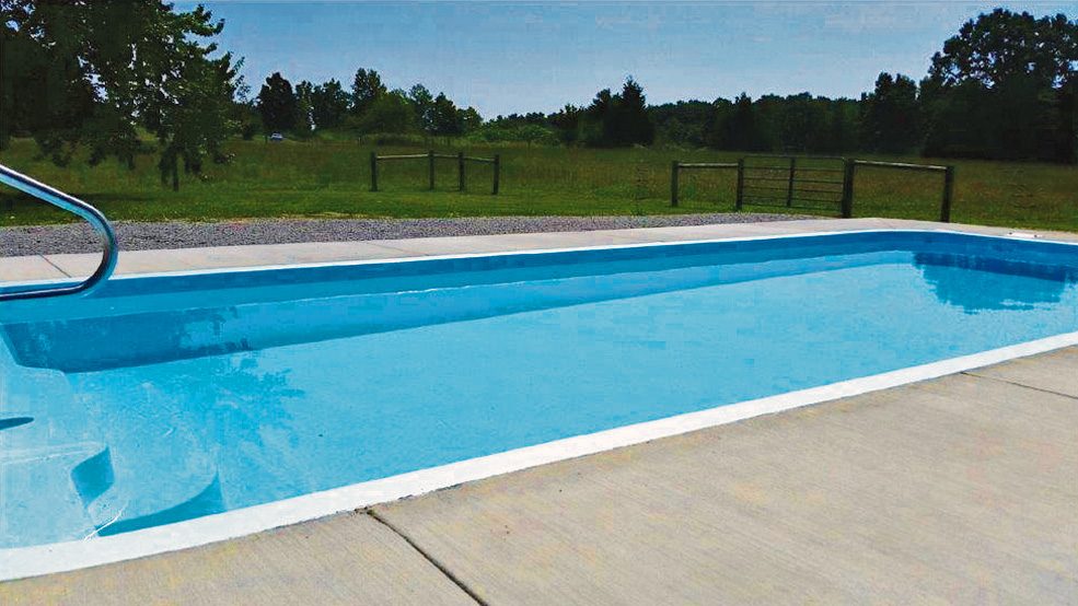 A rectangular vinyl-lined pool with a concrete deck set in a rural yard bordered by trees and a metal fence.