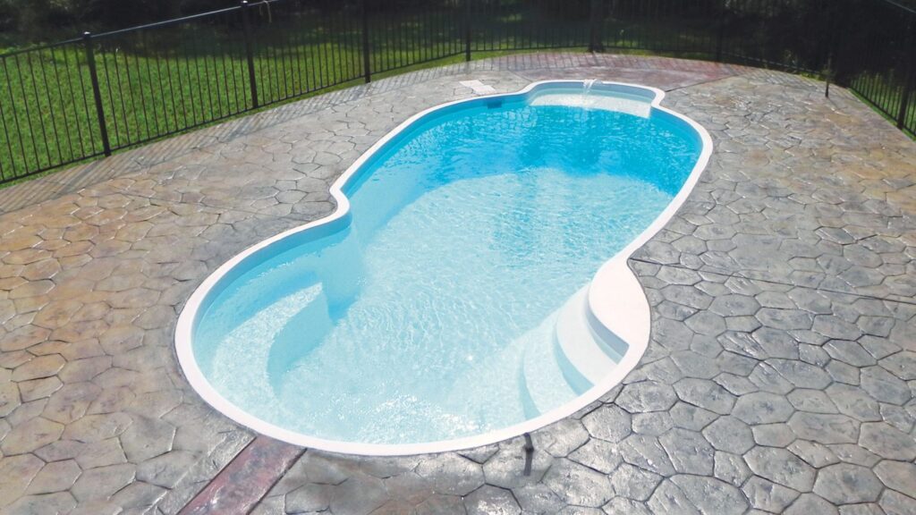 A freeform fiberglass pool with wide steps on a stamped concrete patio bordered by a black metal fence.