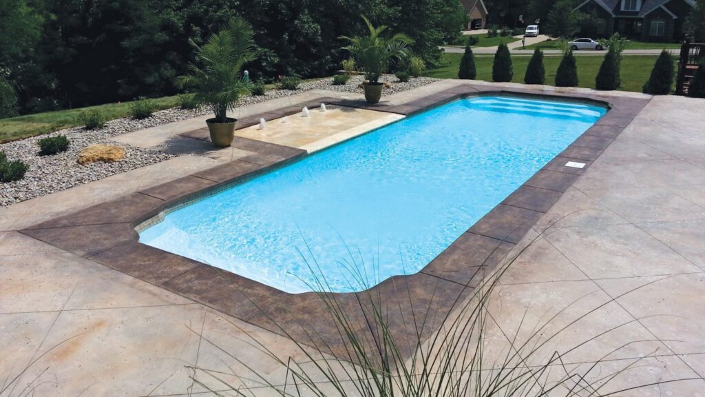 An elevated view of a rectangular fiberglass-style pool with a tanning ledge and stamped concrete decking in a suburban backyard.