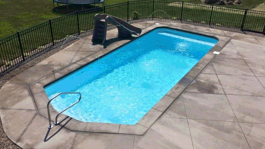 An overhead view of a small rectangular fiberglass pool with a slide and concrete patio surrounded by a black metal fence.