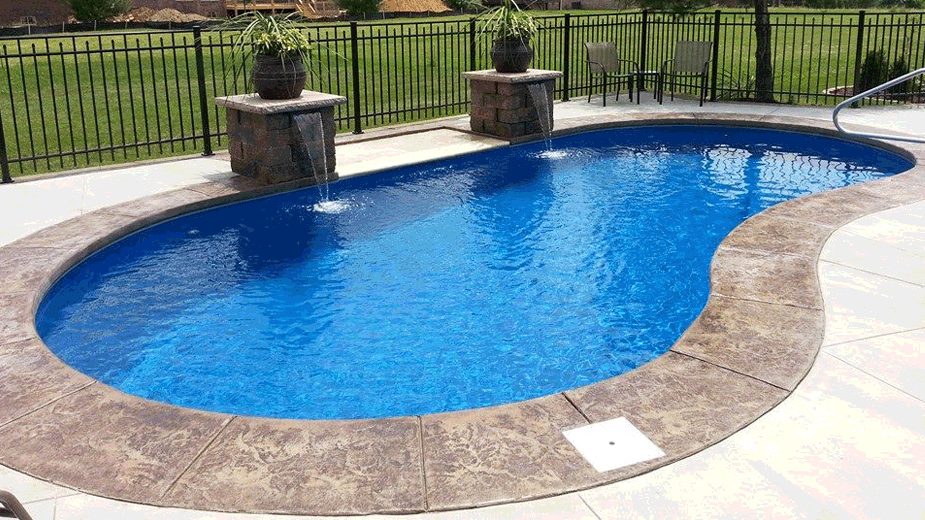A freeform blue fiberglass-style pool with two decorative water spouts on stone pedestals and a stamped concrete patio.