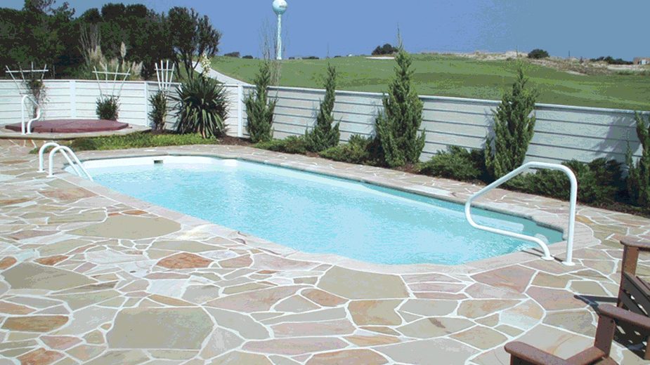 A rectangular pool with rounded ends on a flagstone patio, bordered by a privacy wall and evergreen shrubs.