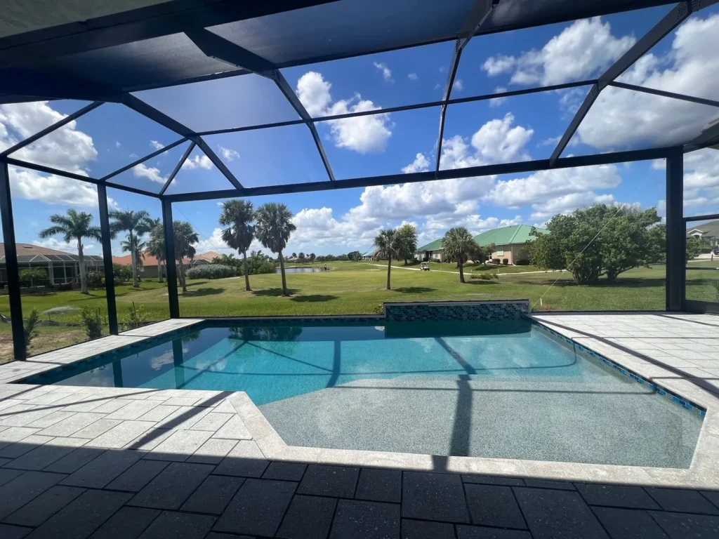 A geometric pool with a raised tiled wall sits under a screened enclosure overlooking a golf course.
