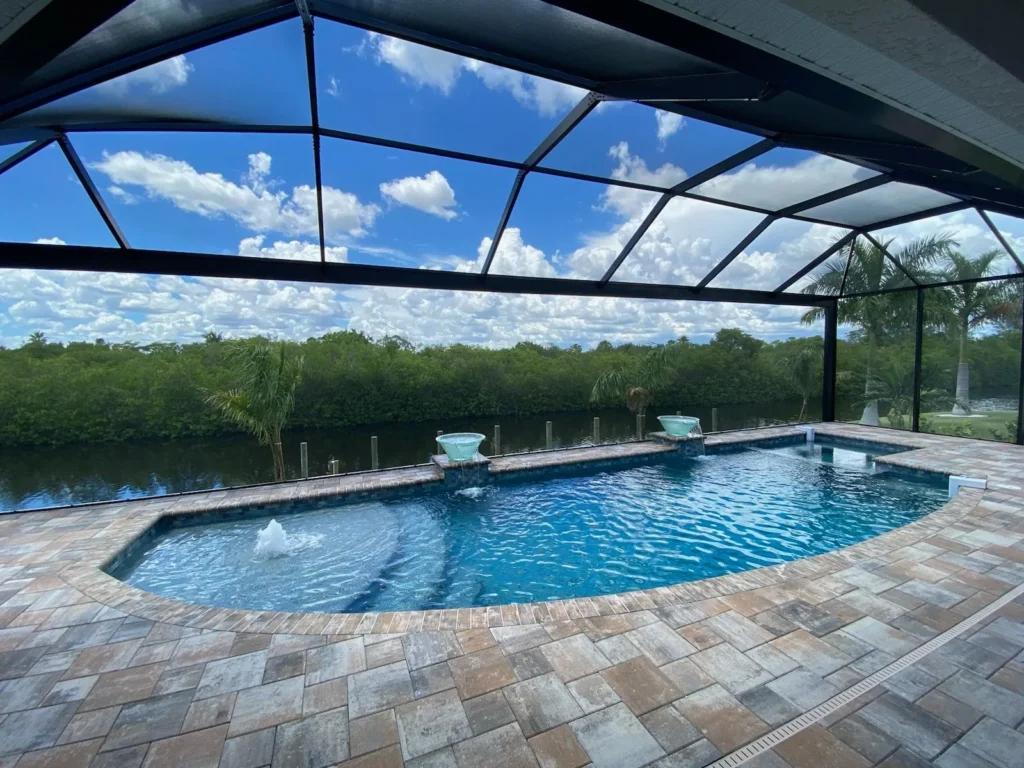A freeform pool with two water bowl fountains and an integrated spa under a screened enclosure overlooking a canal.