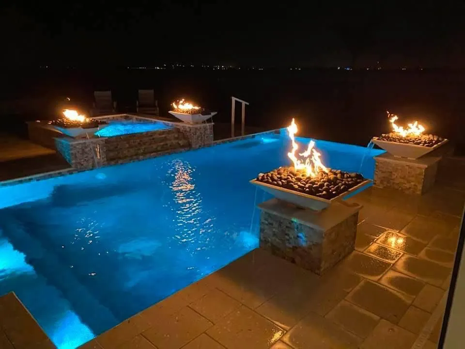 Nighttime infinity-edge pool with a raised spa and four flaming fire bowls spilling water on a waterfront patio.
