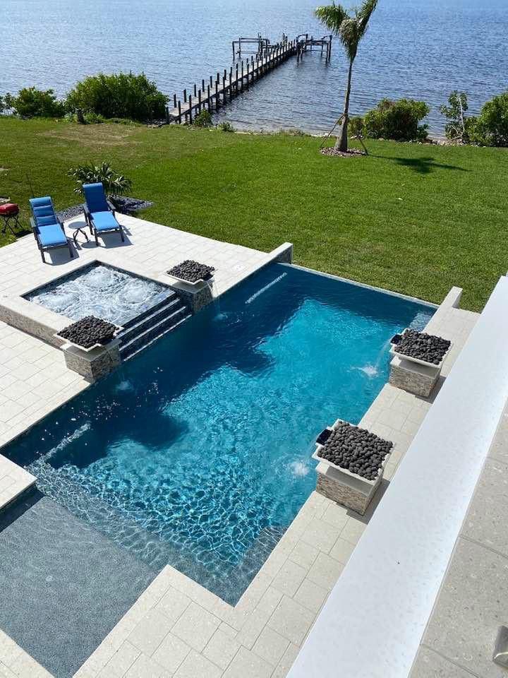 An elevated rectangular pool with a raised spa and stone fire-bowl pedestals overlooking a waterfront dock.