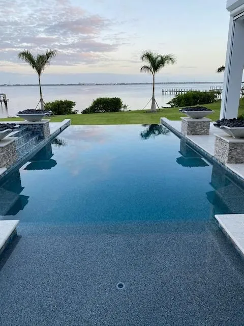 An infinity-edge pool overlooks a wide bay at dusk with palm trees and decorative water bowls on stone pedestals.
