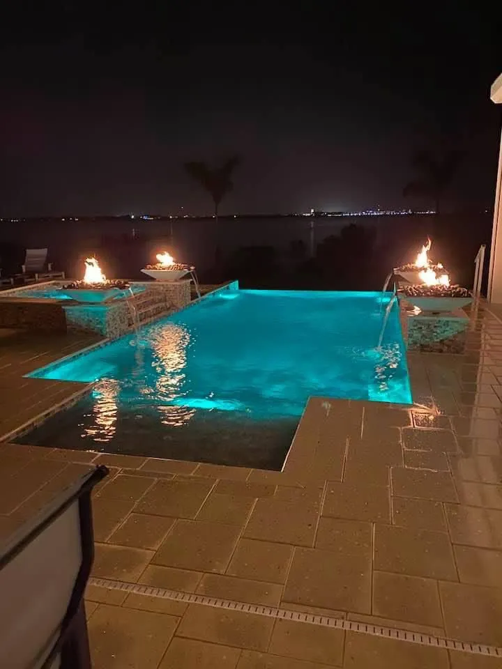 An infinity-edge pool glows turquoise at night with four fire bowls burning on raised pedestals overlooking a dark bay.