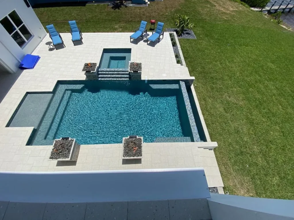 Aerial view of a rectangular backyard pool with a raised spa, tanning ledge, and two fire bowls on a travertine patio.