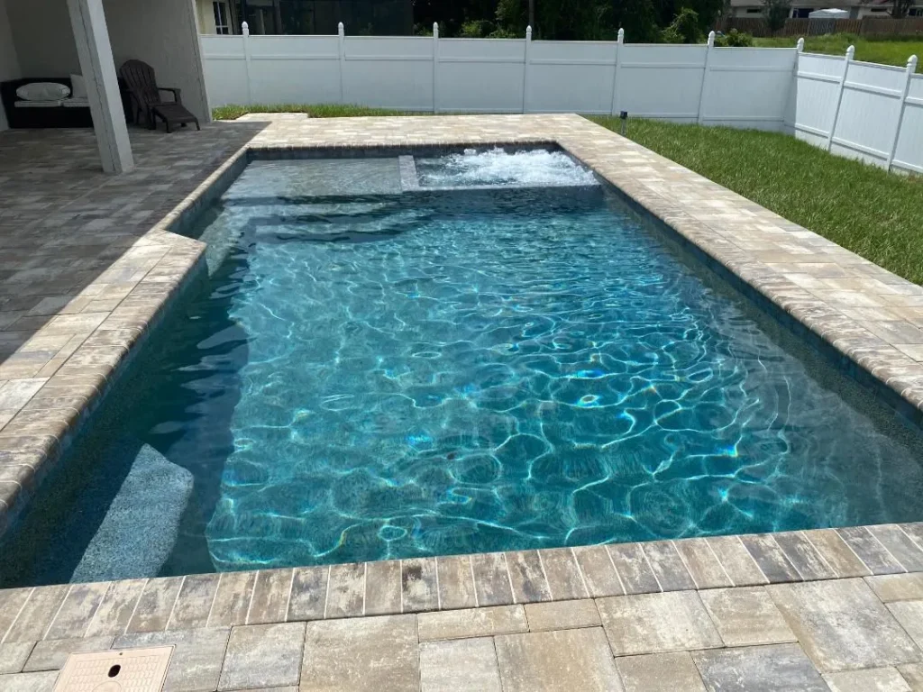 A rectangular backyard pool with an integrated raised spa and jet, surrounded by paver decking and a white vinyl fence.