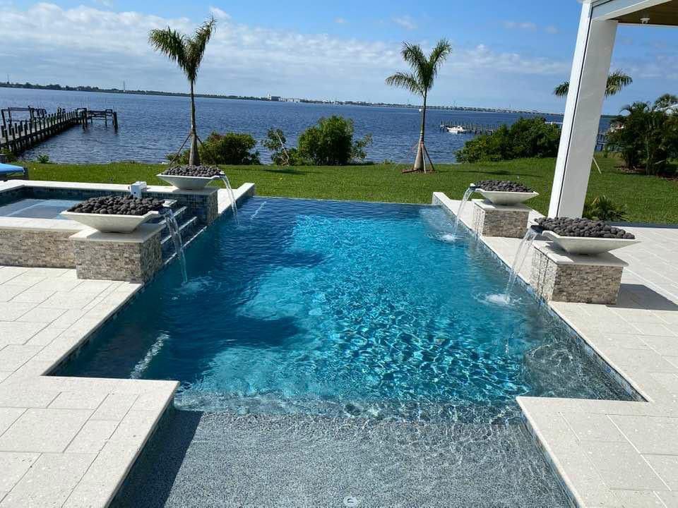 A rectangular pool with a raised spa and four spilling water bowls overlooking a waterfront view with palm trees.
