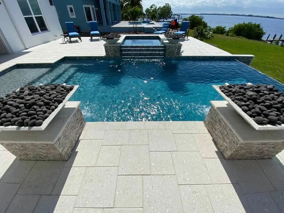 A rectangular pool with a raised spa, spillover water feature, and two lava-rock fire bowls overlooking a waterfront yard.