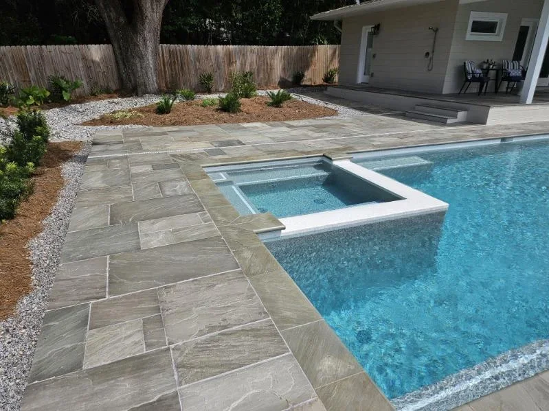 A raised square spa spills into the corner of a pool surrounded by stone paver decking.