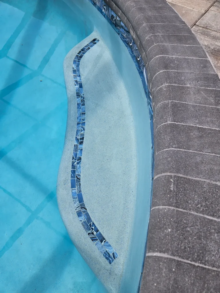 Close-up of a shallow pool shelf edged with a decorative mosaic tile strip beside stone coping.