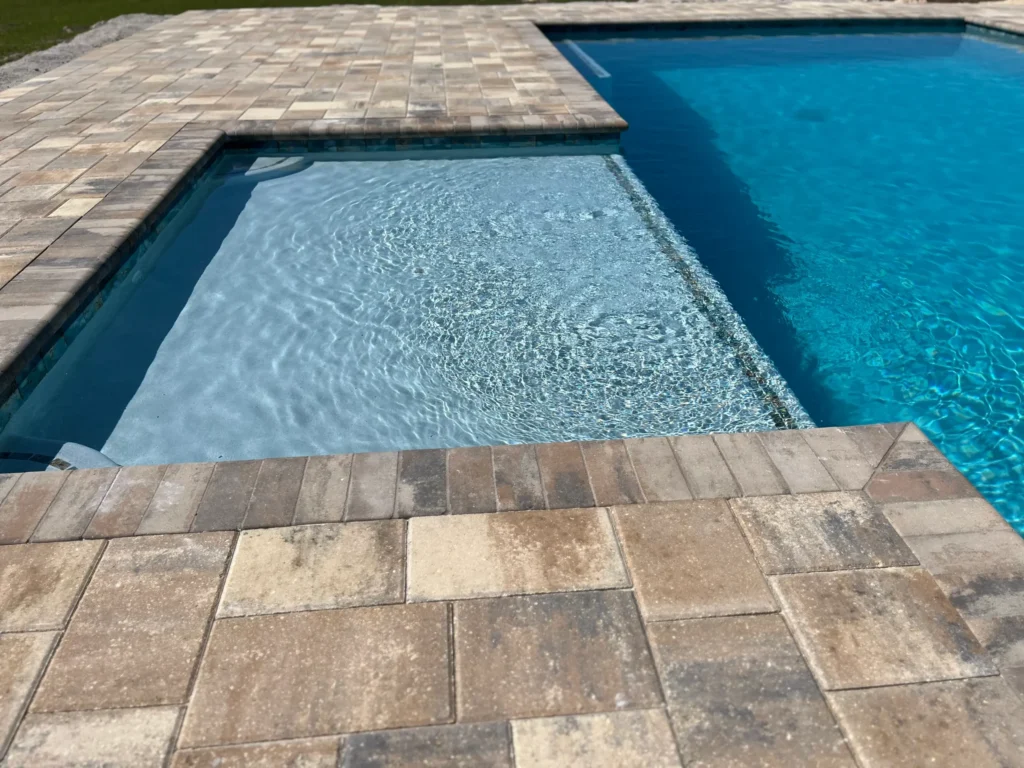 A raised spa spills water into the main pool, seen from a paver deck in the foreground.