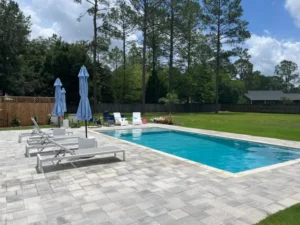 A small rectangular pool with a paver patio, loungers, and umbrellas in a fenced suburban backyard.