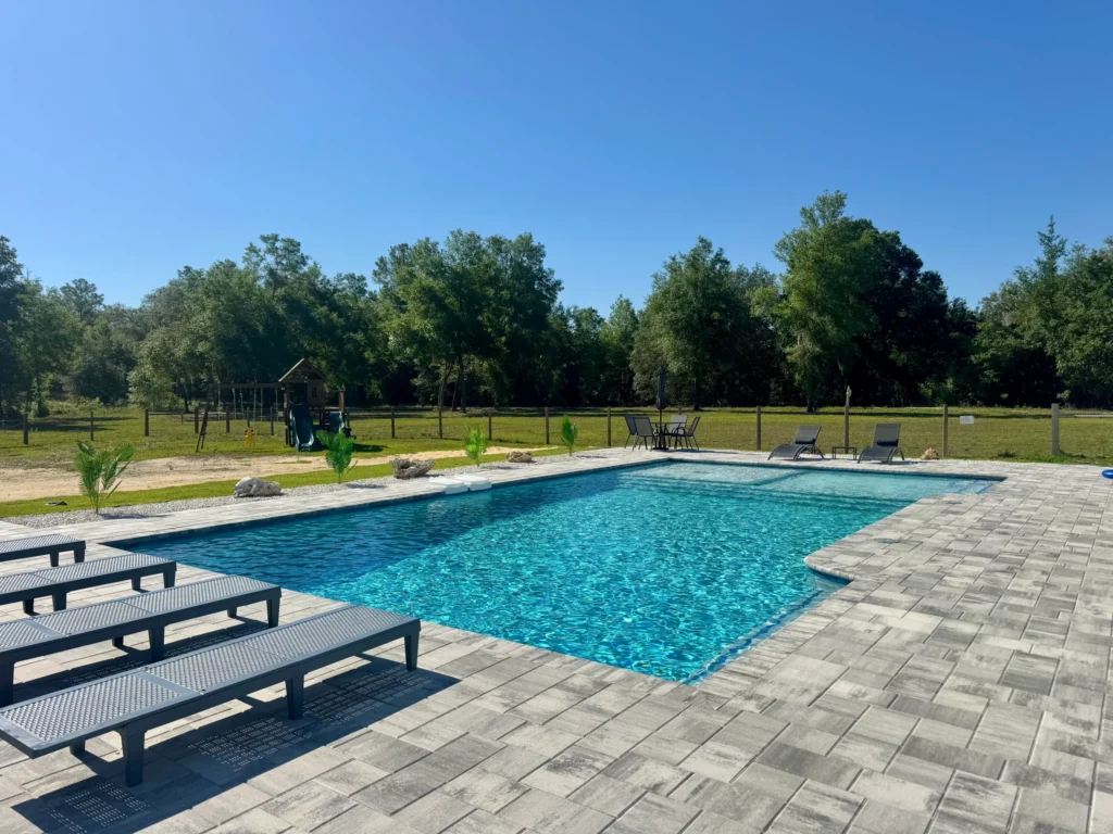 A rectangular backyard pool with a paver deck, loungers, and a play structure in the grassy yard beyond.