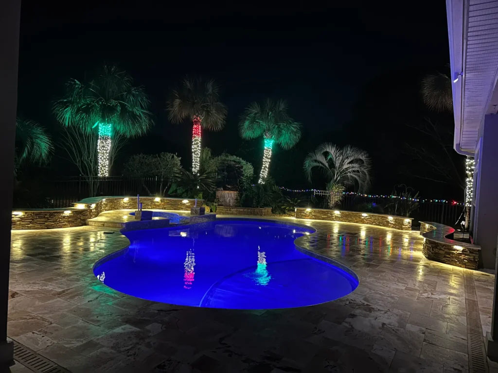 A freeform pool glowing blue at night surrounded by a stone deck and palm trees wrapped in colorful lights.