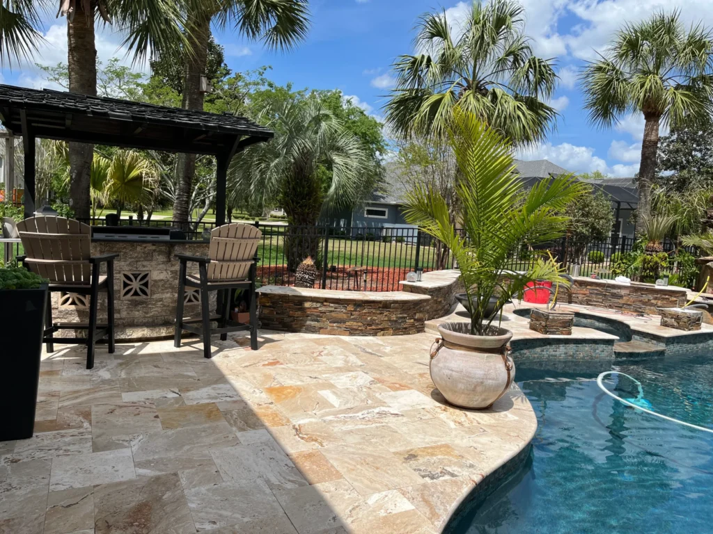 A poolside travertine patio with an outdoor bar and stacked-stone wall, palm trees framing the yard.