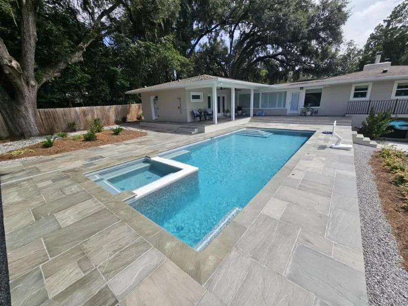 A rectangular backyard pool with a raised spillover spa, surrounded by large gray stone pavers and mature oak trees.