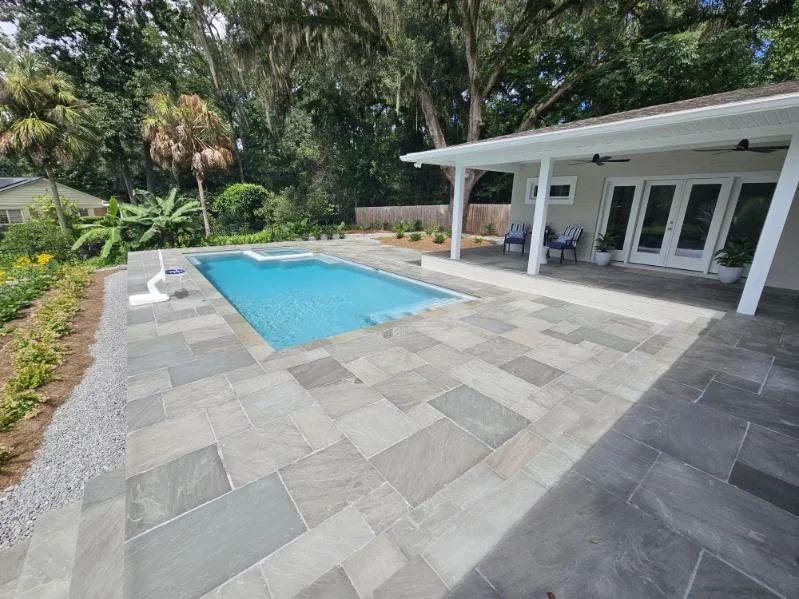 A small rectangular pool with a stone paver patio behind a house with a covered lanai.
