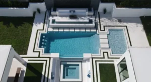 Aerial view of a modern geometric pool with in-water loungers, a raised spa, and water bowl fountains framed by turf and stone patio.