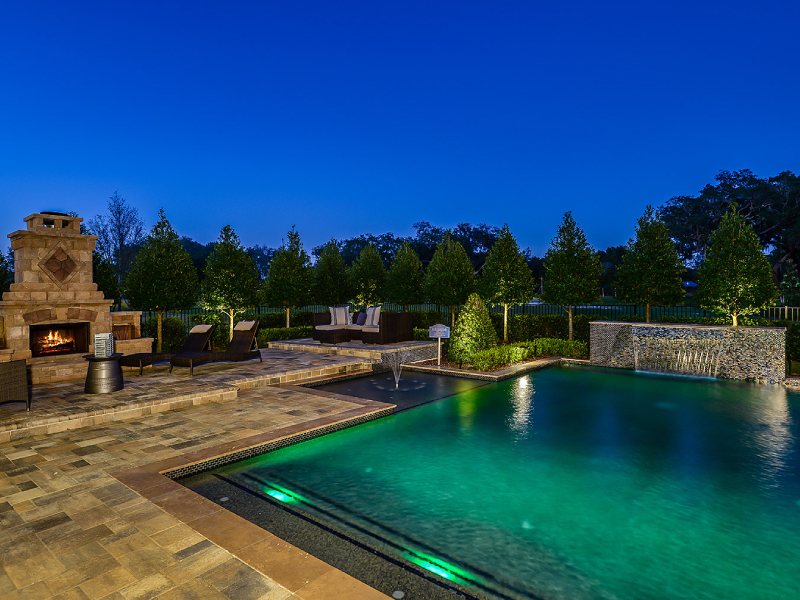 A rectangular pool glows turquoise at dusk beside a stacked-stone waterfall wall and an outdoor fireplace with lounge seating.