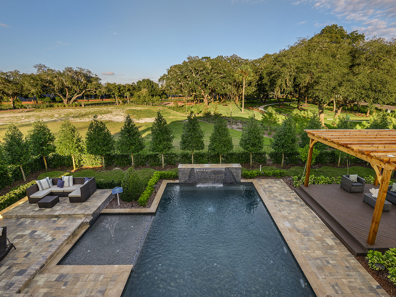 Rectangular pool with a stone waterfall wall and separate fountain feature, bordered by a paver patio, pergola, and open green fairway with mature oaks.