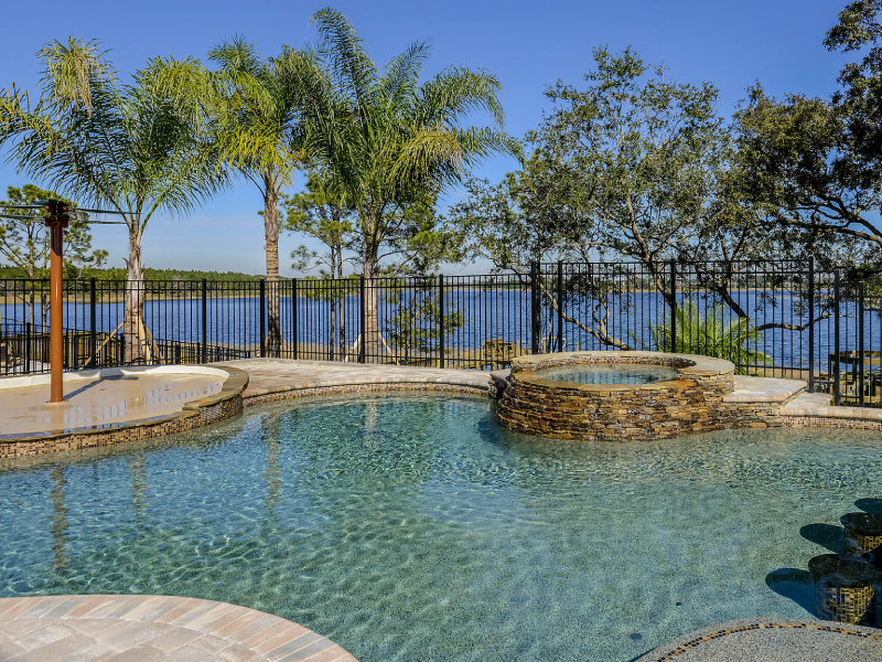 A freeform pool with a raised stone-clad spa overlooks a lake behind a black metal fence.