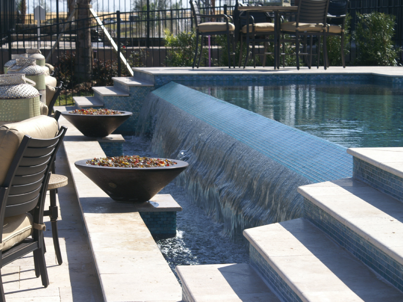 A tiled water wall cascades into a pool beside two large decorative bowls on a travertine patio.