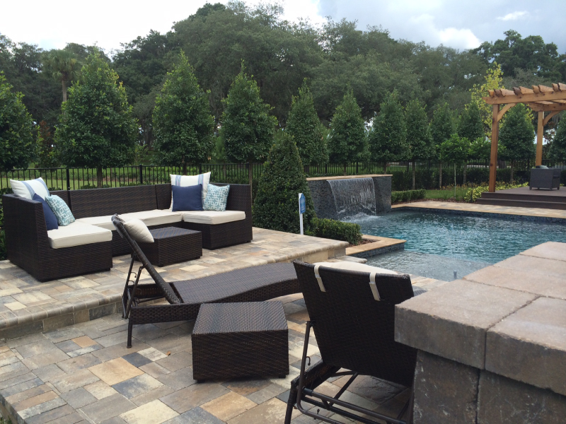 Outdoor lounge seating on a paver patio with a pool and raised water feature wall in the background.