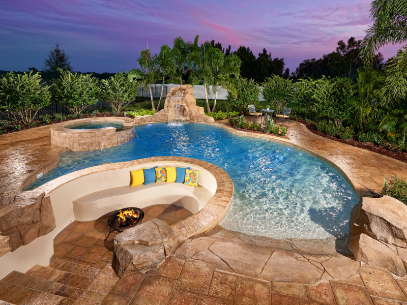 A freeform backyard pool with a raised spa, rock waterfall slide, and curved fire pit seating area at dusk.