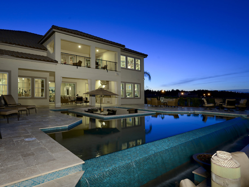 A large infinity-edge pool with a central seating island glows at dusk beside a grand two-story home.
