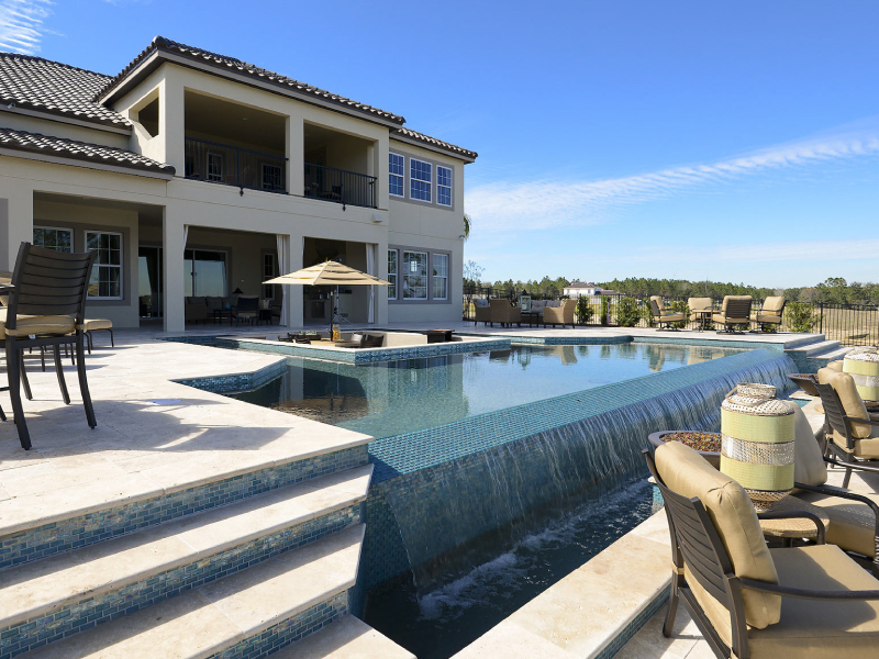 A luxury infinity-edge pool with a tiled waterfall wall behind a large two-story home under a clear blue sky.