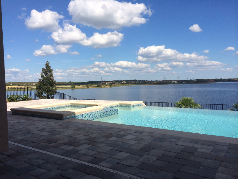 An infinity-edge pool with a raised tiled spa overlooks a large lake under a blue sky.