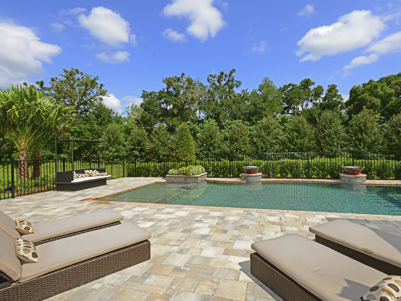 A rectangular backyard pool with two spilling water bowls on pedestals, framed by a paver patio, loungers, and a wooded tree line beyond a black metal fence.