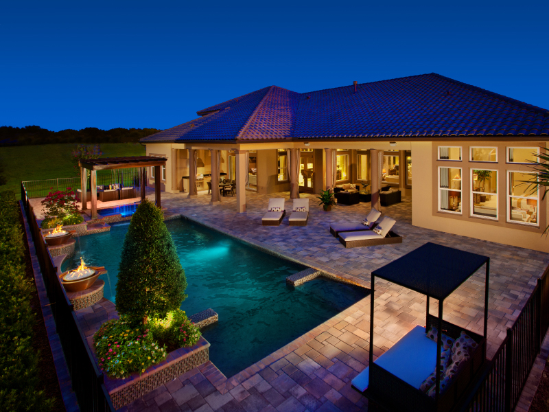 Twilight view of an elegant geometric pool with fire bowls and a paver patio behind a large tile-roofed home.