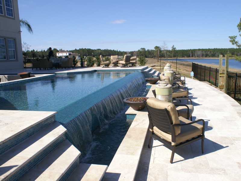 An elevated rectangular pool with a glass-tile knife-edge waterfall spilling into a lower catch basin beside a lakefront patio.